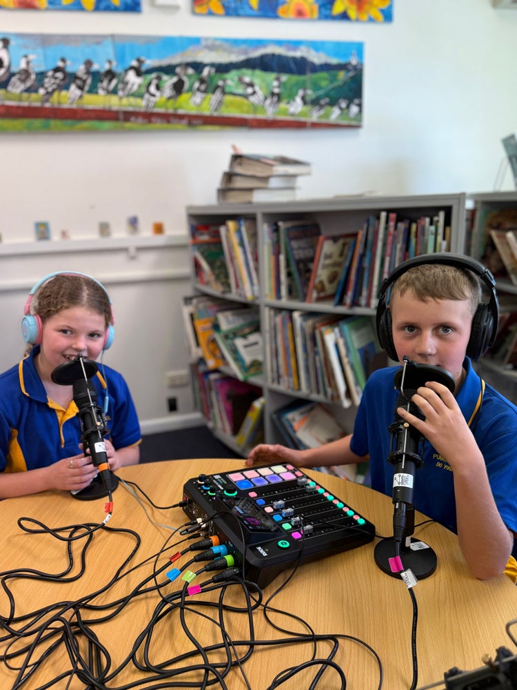 Photo of two students sitting at a round table with headphones on and talking into microphones