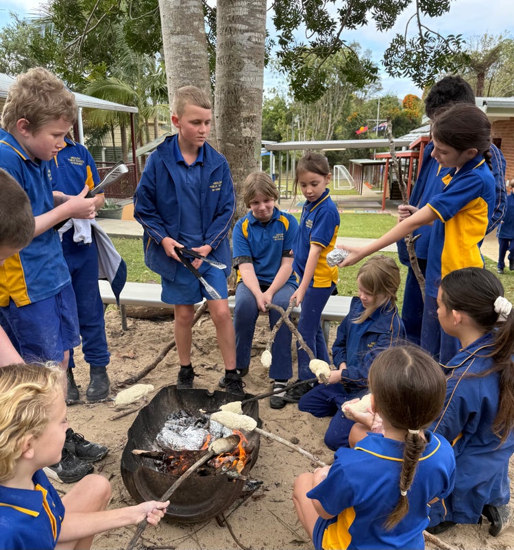 Photo of a group of ten students standing and sitting around a small fire and toasting dampers